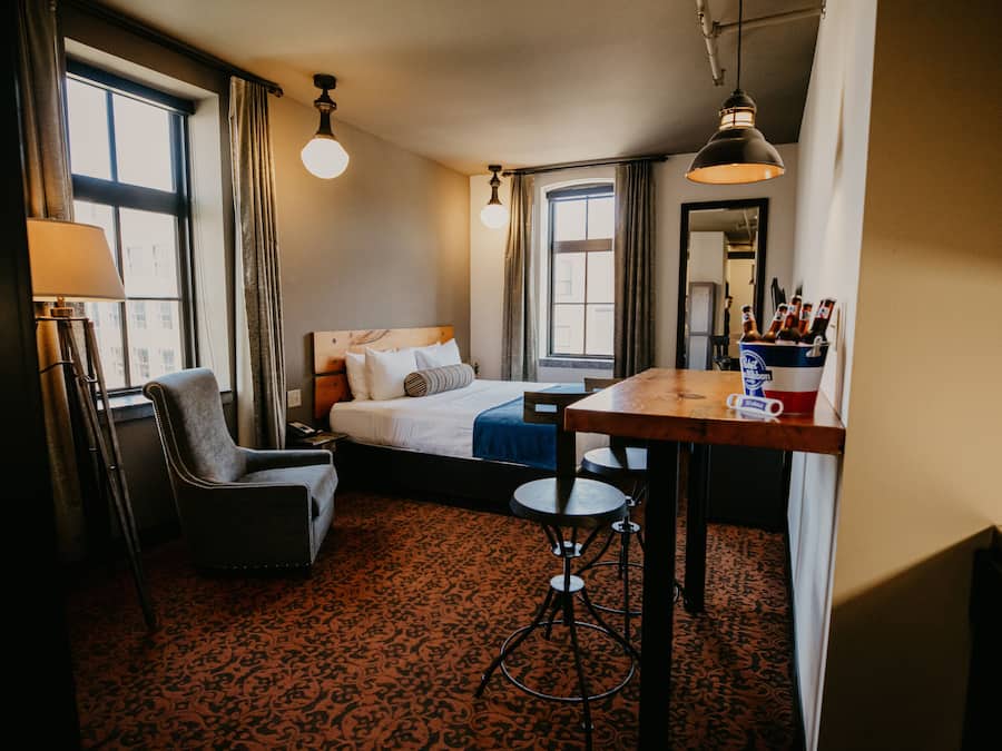 A hotel room with a bed, a desk, a chair, a lamp, and a window.