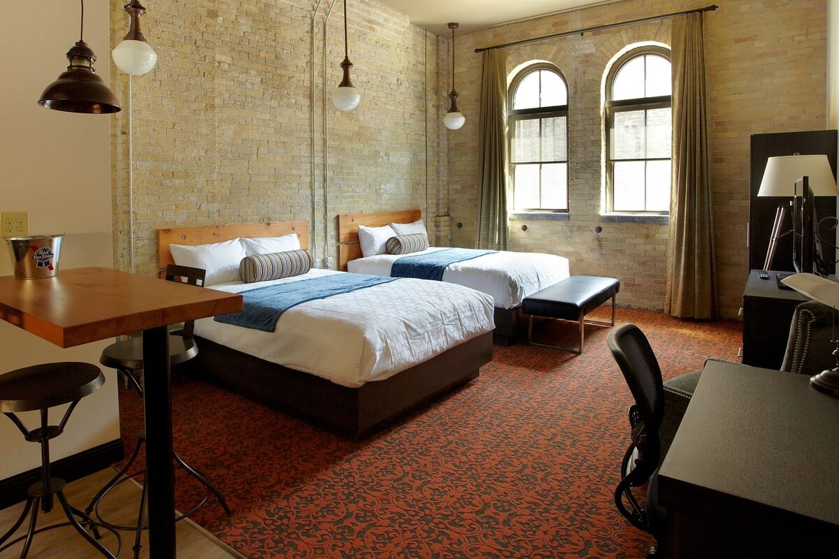 A hotel room with two beds, a desk, a chair, and a table. There are two windows with curtains, a brick wall, and hanging lamps.