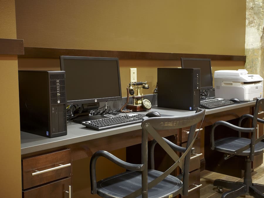 A hotel workspace with three computer stations, each with a monitor, keyboard, and mouse. There is a printer on the right side of the desk.