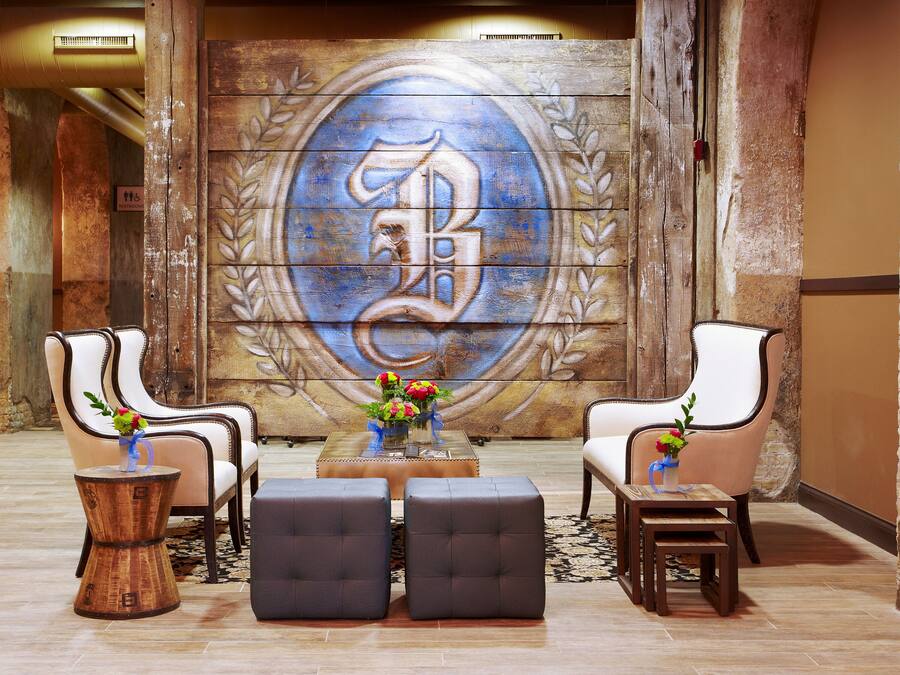 Een hal met twee fauteuils, een bijzettafeltje en poefs. Een groot muurschilderij met een monogram en laurierkrans is zichtbaar.
