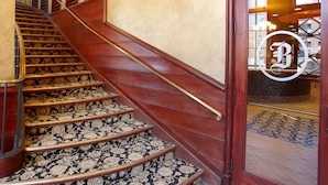 A grand staircase with ornate carpet and wooden railings leading to a door with a logo. - The Brewhouse Inn & Suites (Milwaukee)
