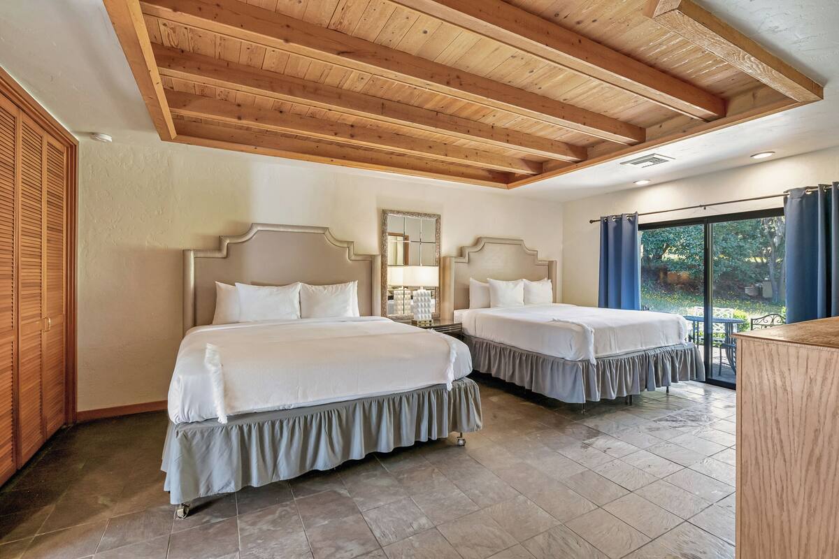 A hotel room with two beds, wooden ceiling beams, a large window with curtains, and a view of greenery outside.