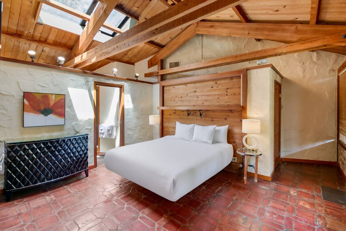 A bedroom with a large bed, wooden beams, a stone fireplace, and a view through a glass door.