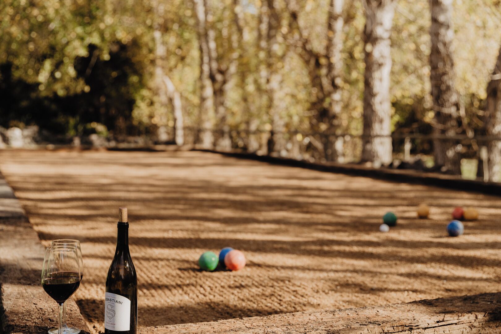 A bottle of wine and a glass on a wooden table in a park with a croquet field in the background.