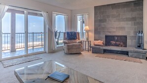 A spacious living room with a glass coffee table, a comfortable armchair, a fireplace, and large sliding glass doors leading to a balcony. - Cavalier Beachfront Condominiums (Lincoln City)