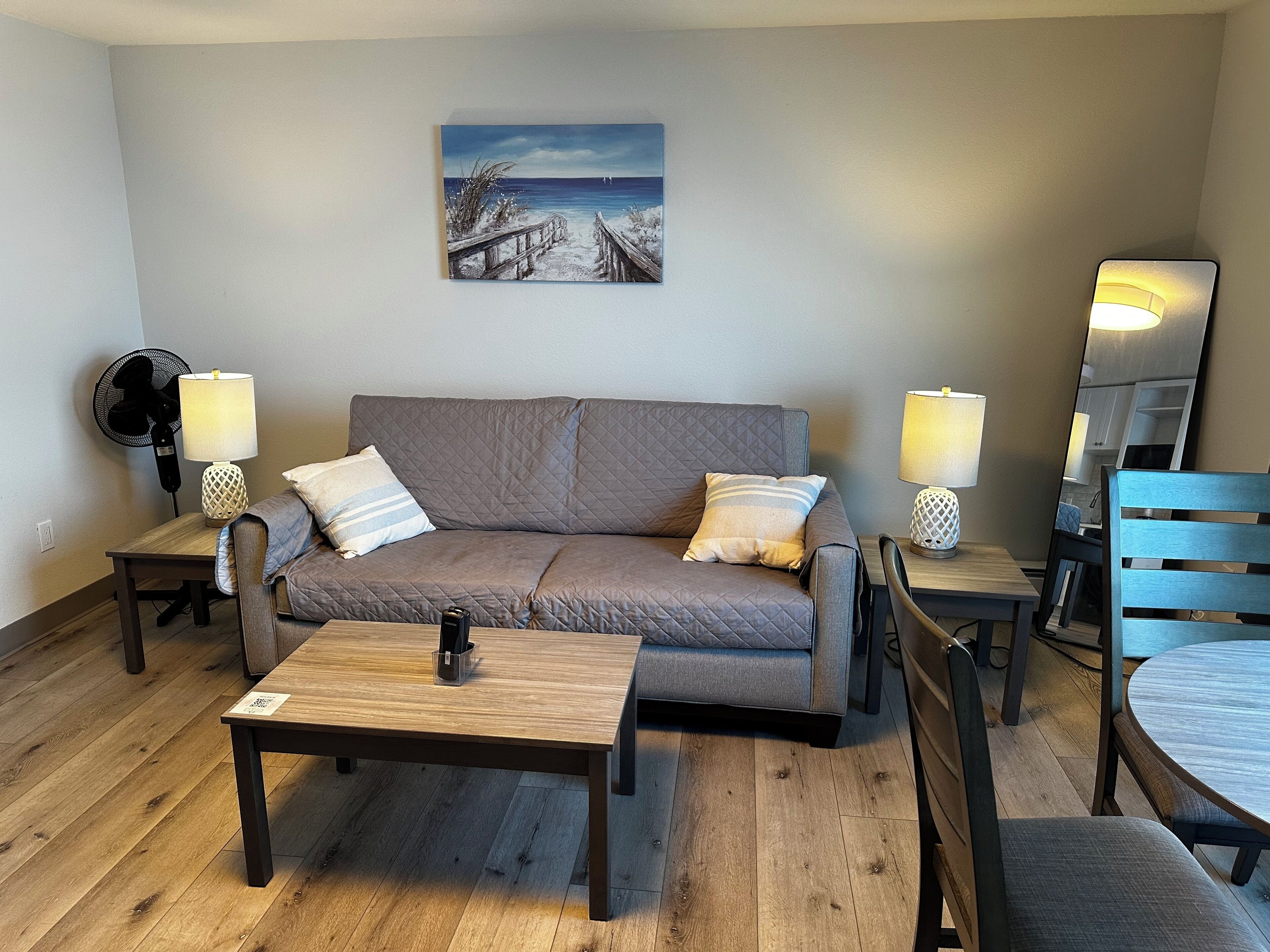 A cozy living room with a sofa, two side tables with lamps, a coffee table, a fan, and a painting on the wall.