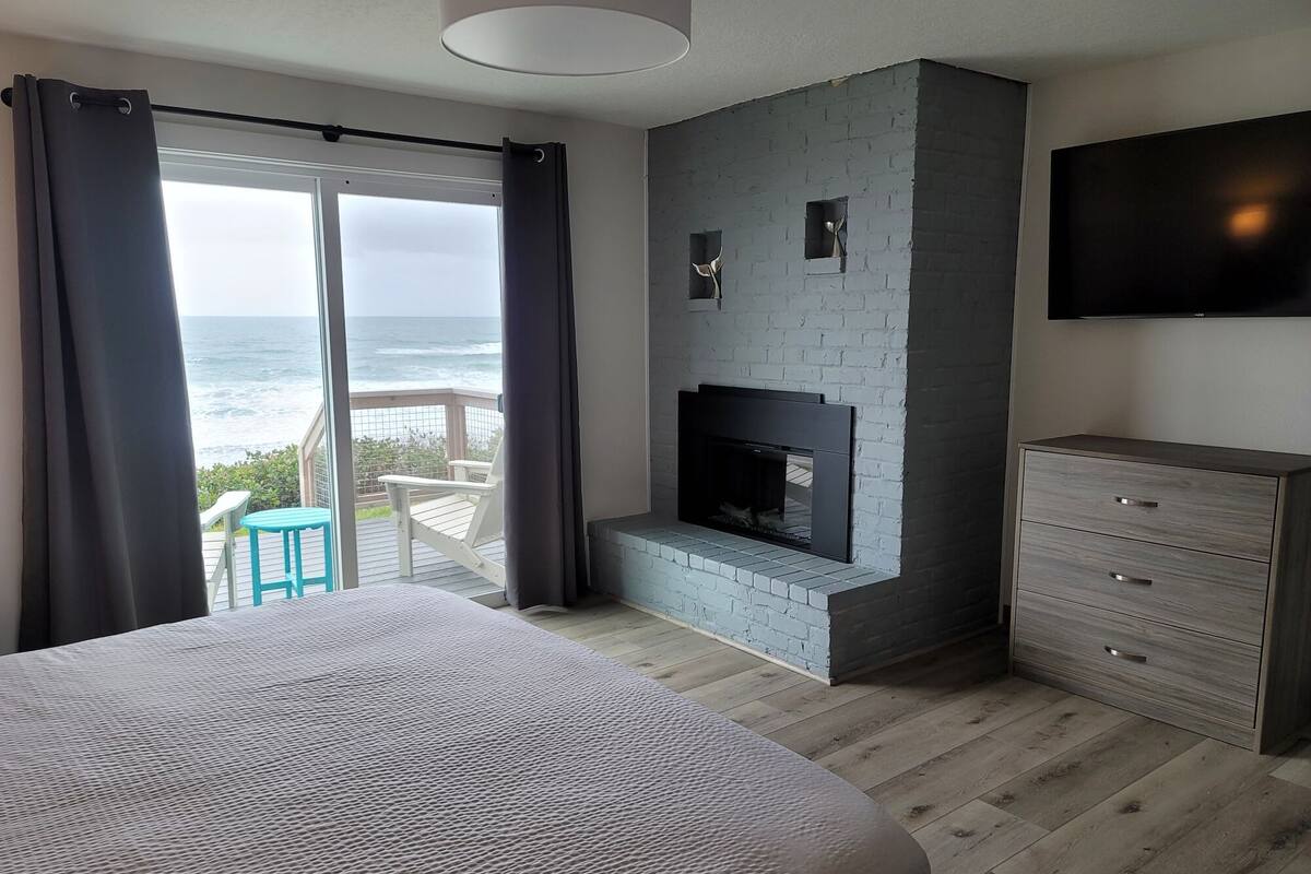 A bedroom with a large bed, a fireplace, a TV, and a view of the ocean.