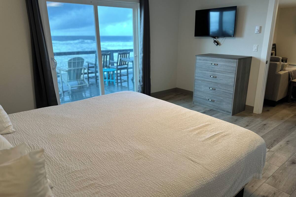 A bedroom with a bed, a dresser, and a TV. There is a view of the ocean through a sliding door.
