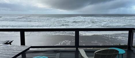 A deck with two Adirondack chairs and a small table, overlooking a beach with rough waves under a cloudy sky.