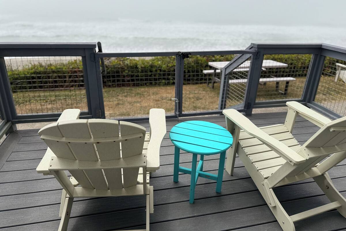 A deck with Adirondack chairs and a small table, overlooking a grassy area with a fence and picnic tables in the background.
