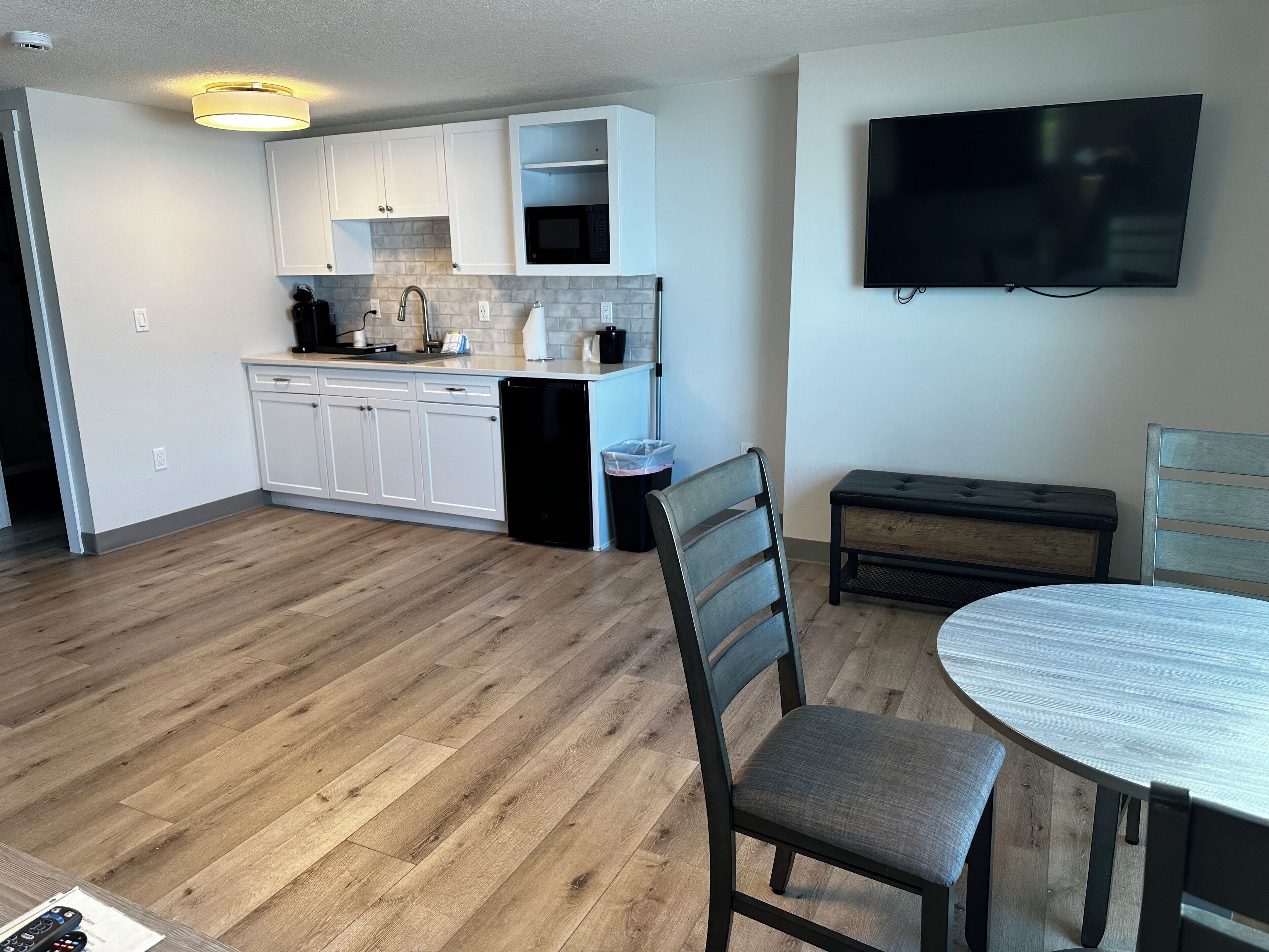 A modern kitchen with white cabinets, a microwave, and a dishwasher. There is a dining area with a round table and two chairs. A flat-screen TV is mounted on the wall.