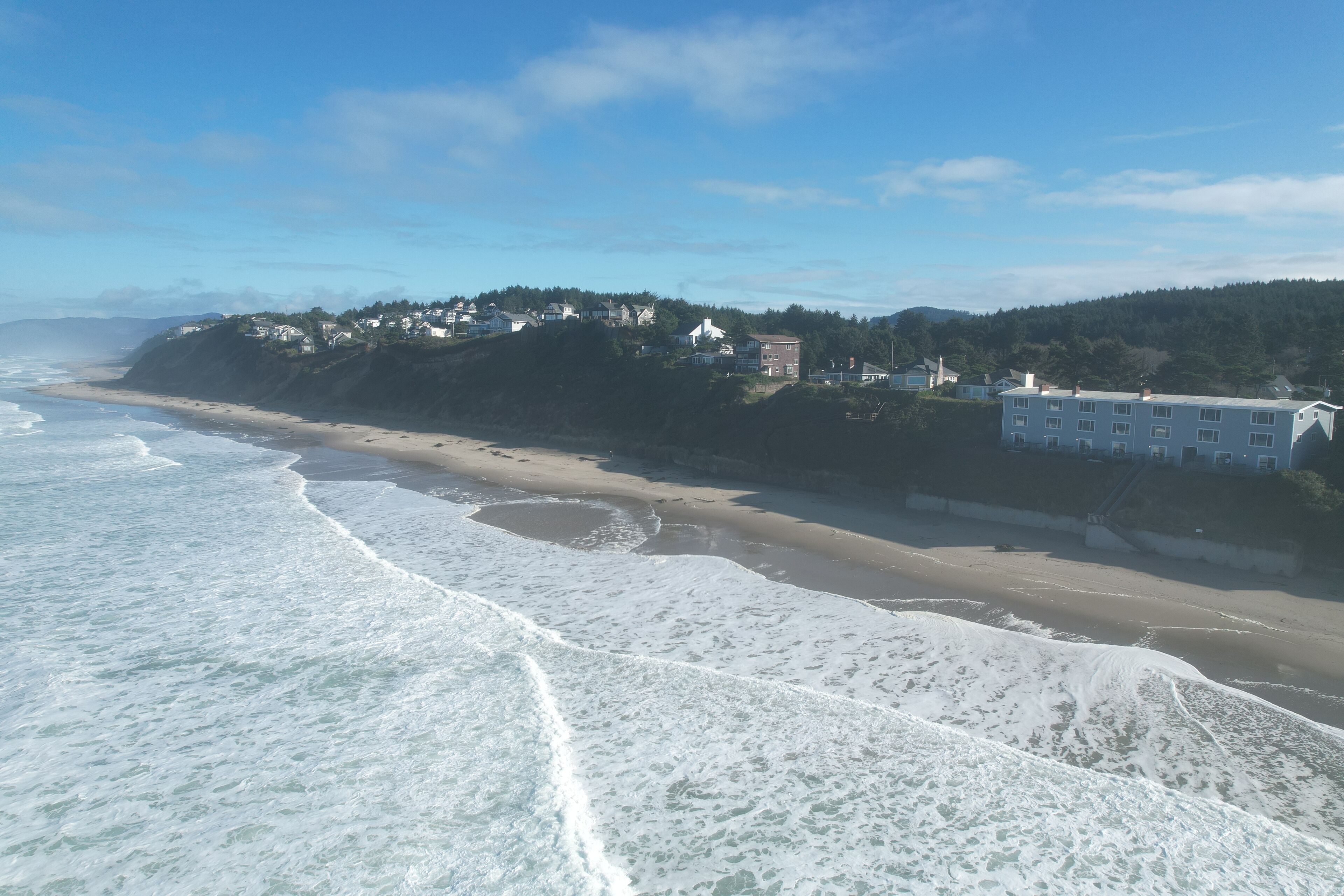 A coastal view with a sandy beach, ocean waves, and a hillside with buildings.