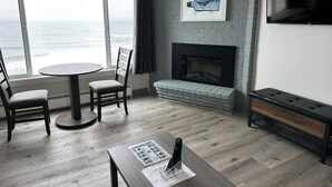 A modern living room with a fireplace, a TV mounted on the wall, and a view of the ocean through large windows. - West Beach Suites (Lincoln City)