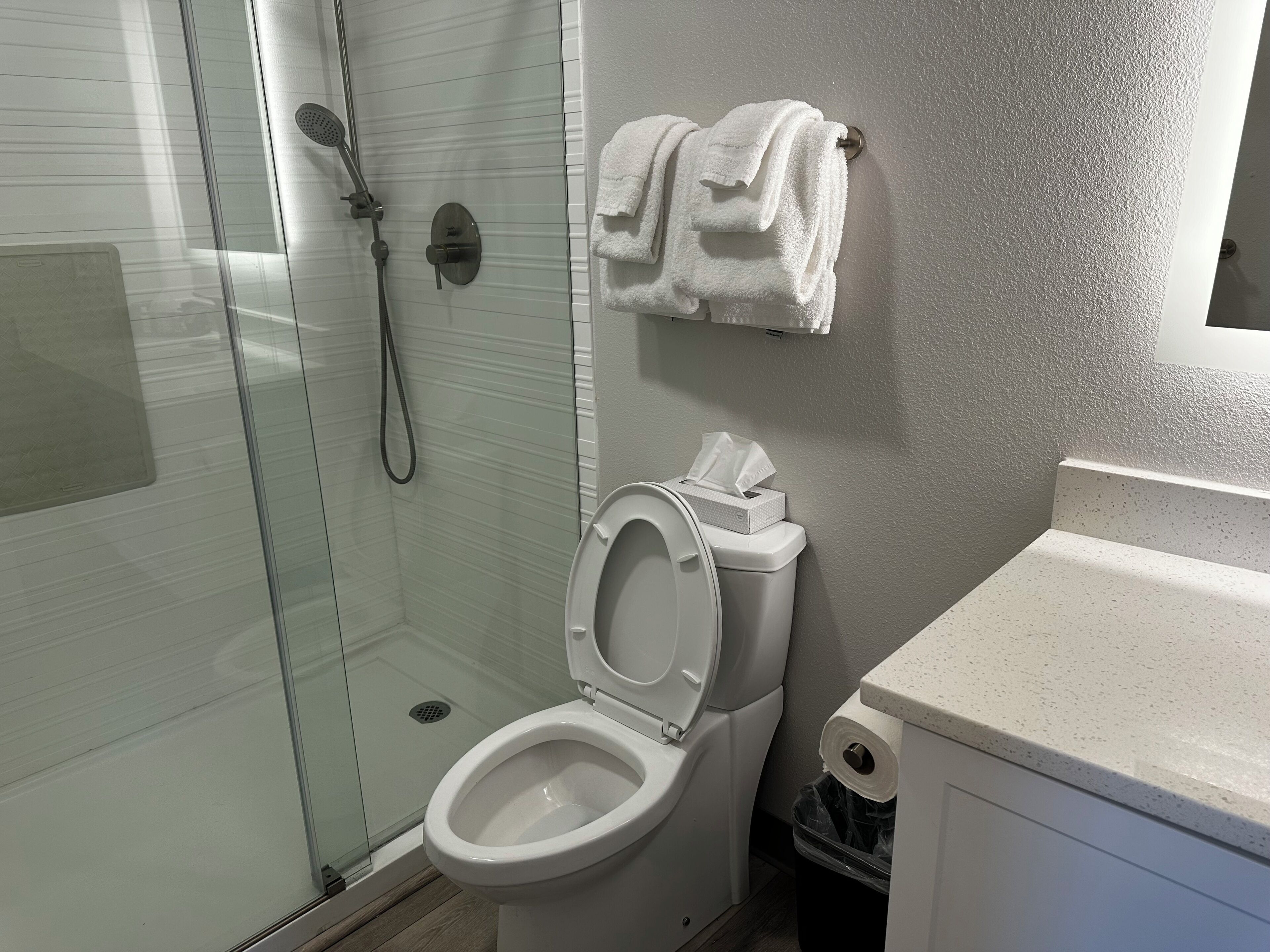 A bathroom with a glass-enclosed shower, a toilet with a paper towel dispenser, and a towel rack with folded towels.