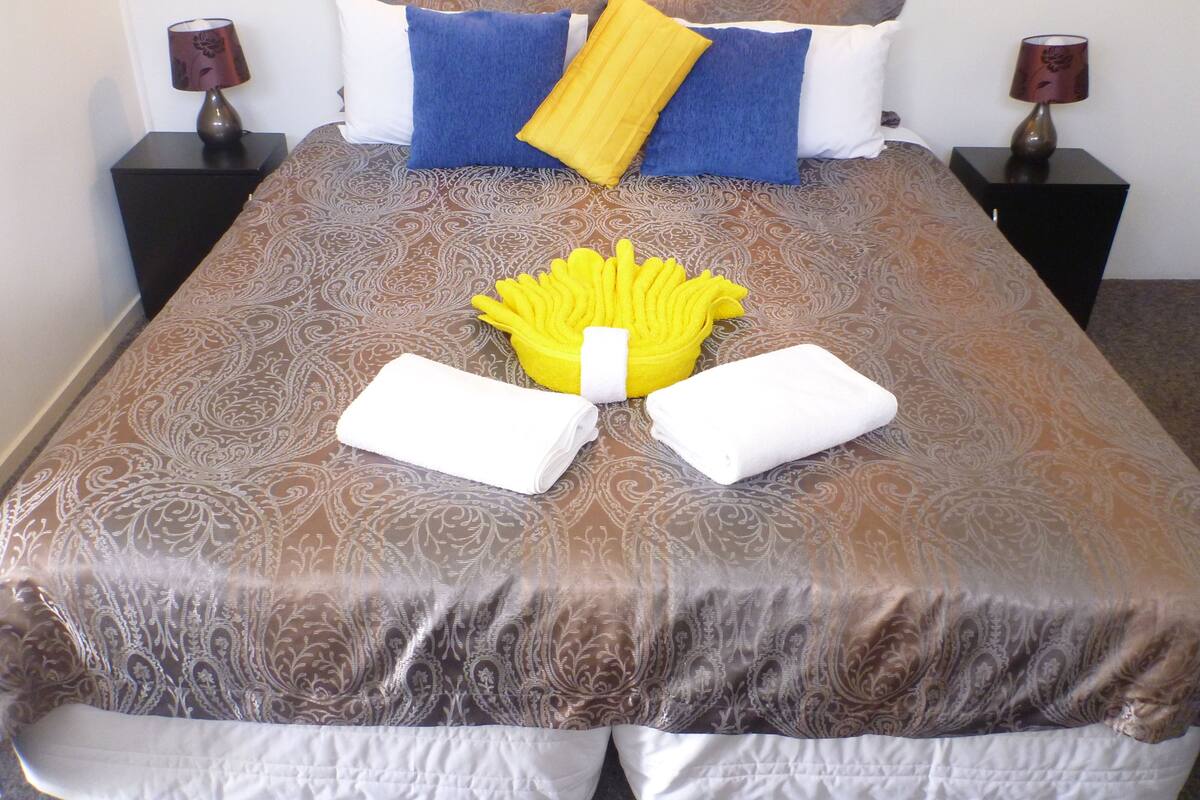 A neatly made bed with a patterned bedspread, two pillows, a yellow towel, and two folded towels.