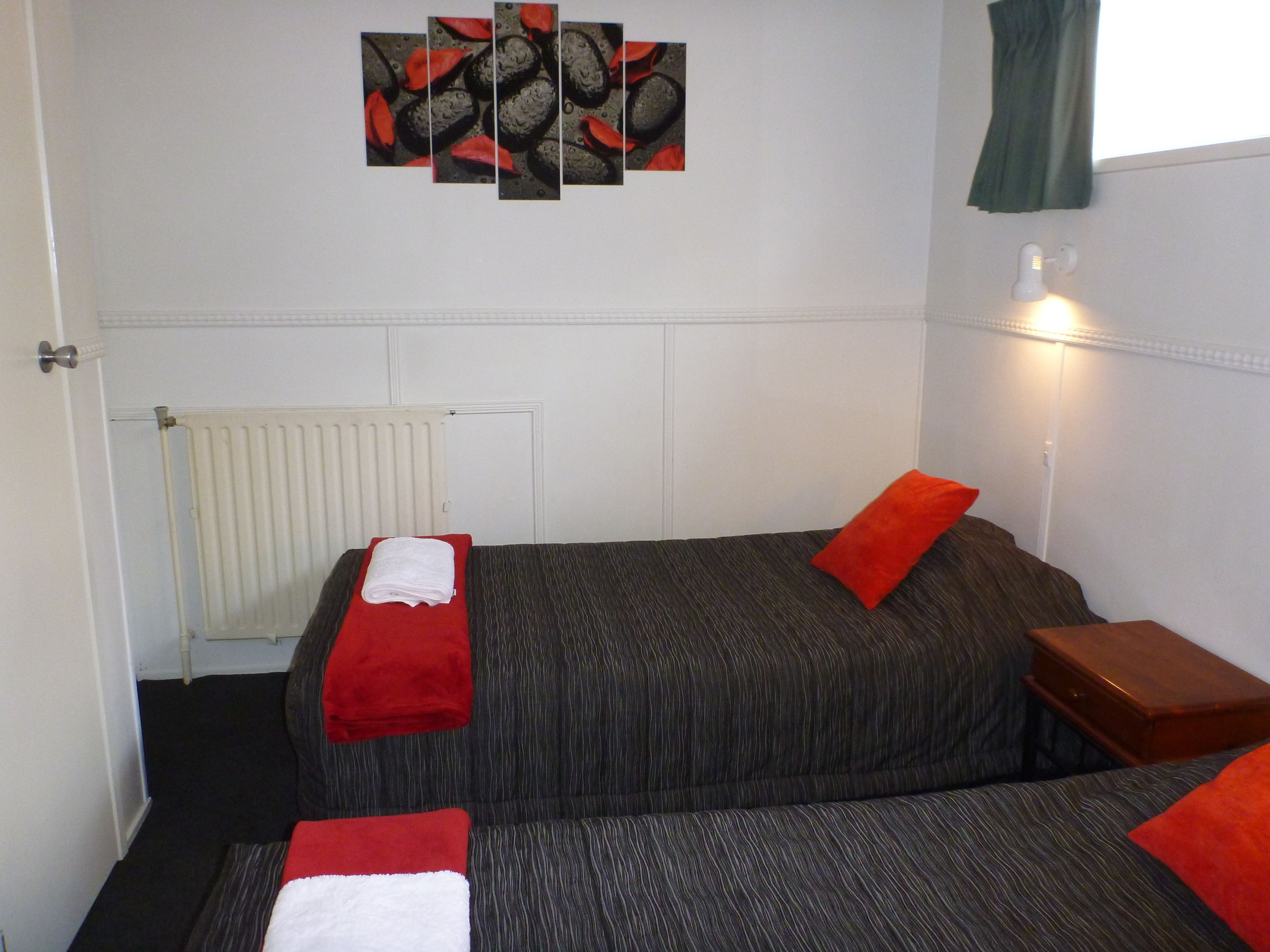 A hotel room with a bed, a red pillow, a white towel, a radiator, and a wall art piece.