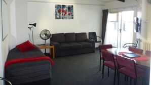 A living room with a sofa, a dining table with chairs, a fan, and a painting on the wall. - Fernleaf Motel (Rotorua)
