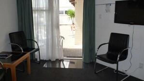 A room with two black chairs, a wooden table, a TV mounted on the wall, and a window with green curtains. - Fernleaf Motel (Rotorua)