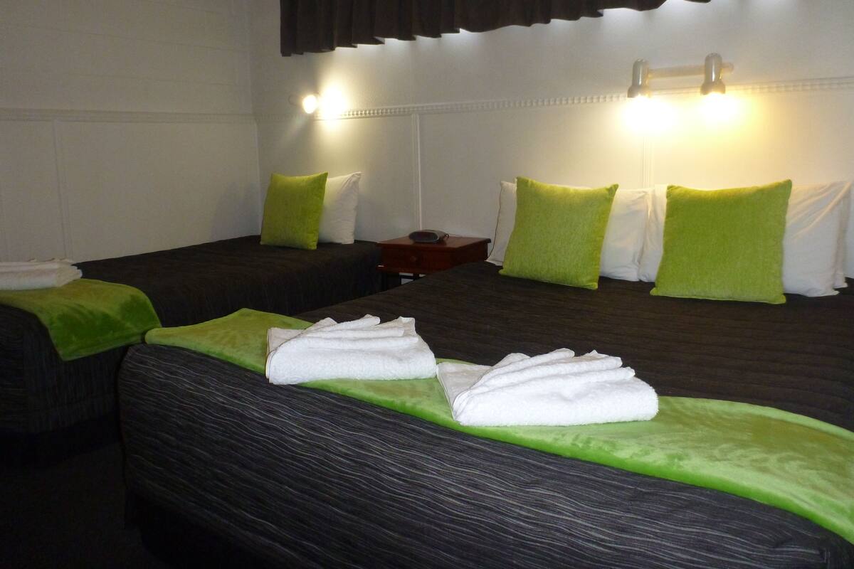 A hotel room with two beds, each with green and white pillows, and a nightstand with a lamp.