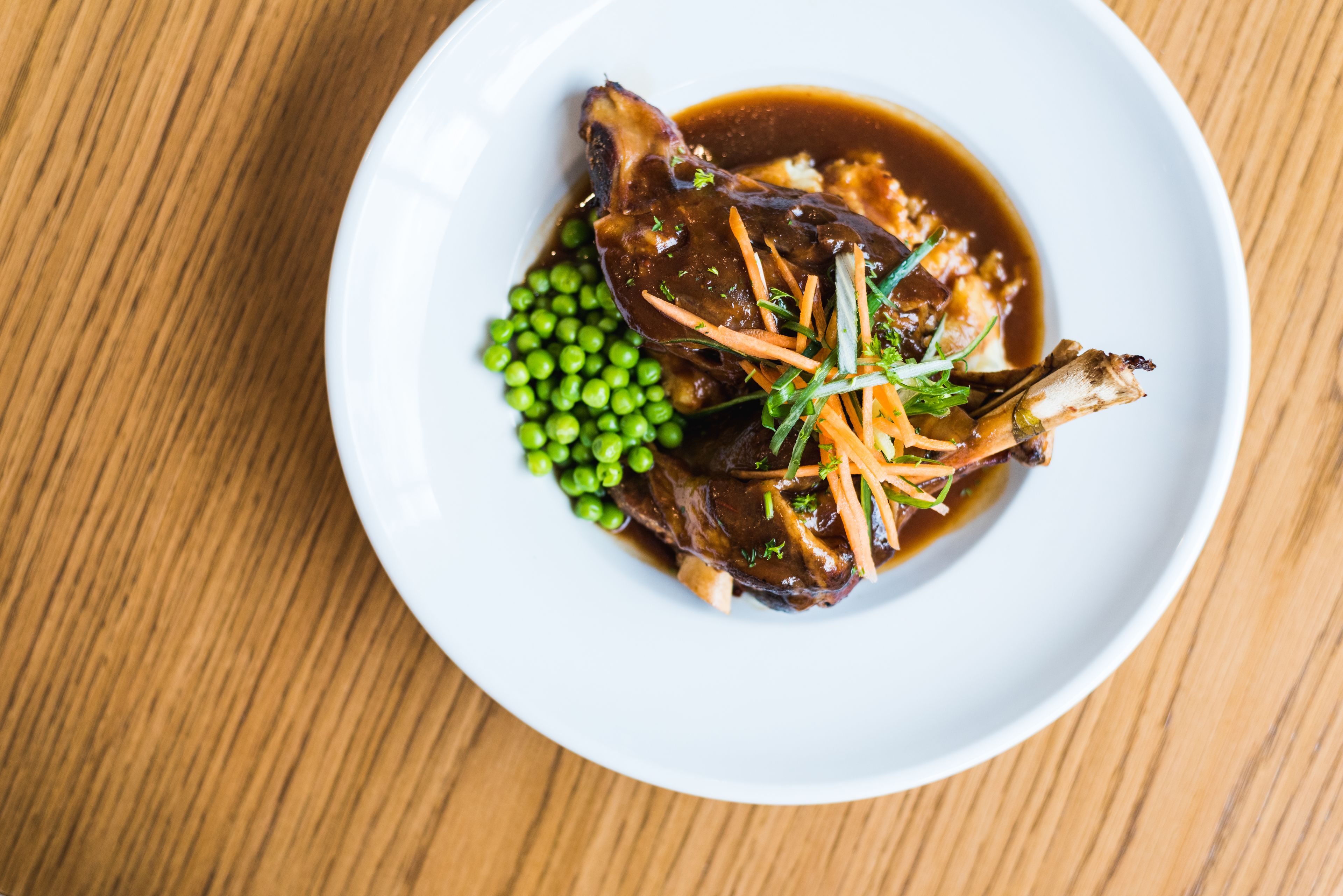Une assiette blanche contenant un plat de viande, des petits pois verts et des carottes, posée sur une table en bois.
