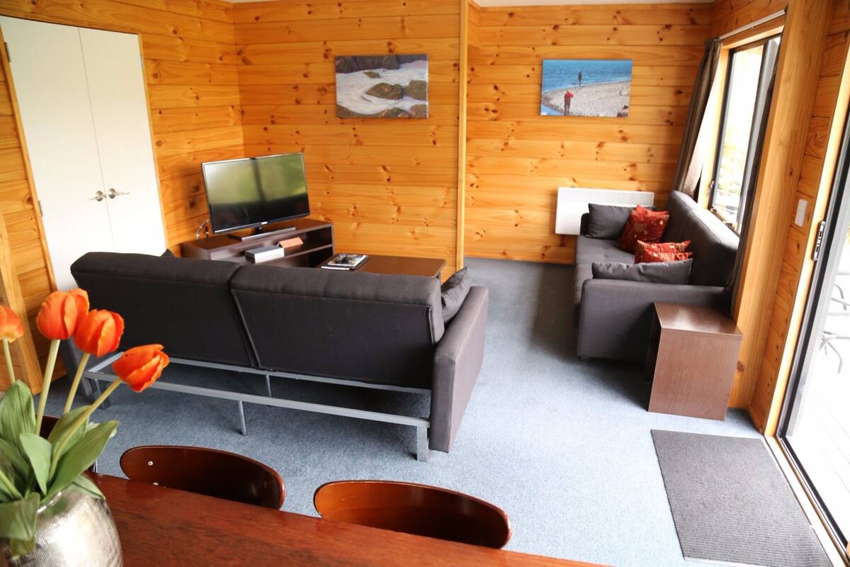 Interior de una cabaña de madera con sofá, televisión y mesa de comedor.