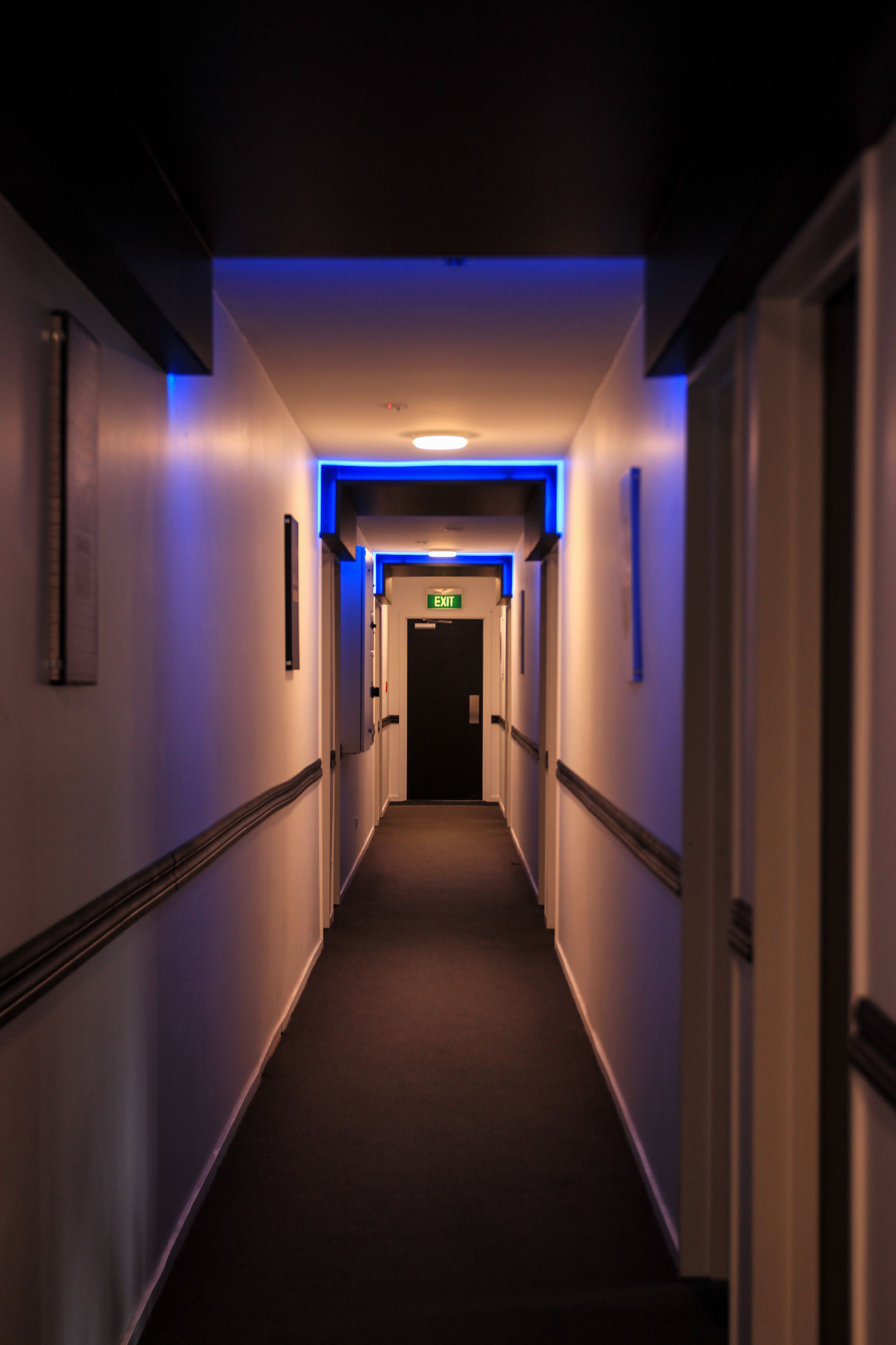 A narrow, dimly lit hallway with a blue accent lighting on the ceiling, leading to an exit door.