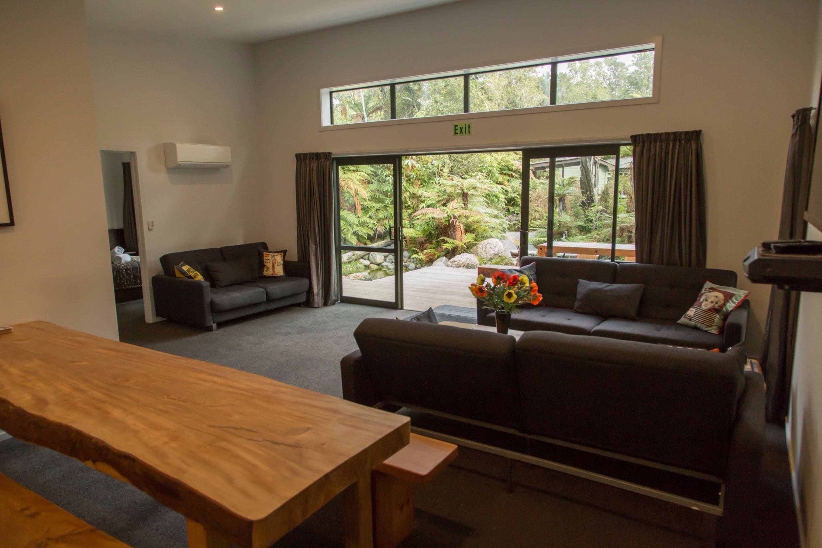 Uma sala de estar moderna com uma mesa de madeira, um sofá e um divã. Há uma janela grande com cortinas, um aparelho de ar condicionado e uma placa indicando uma saída.
