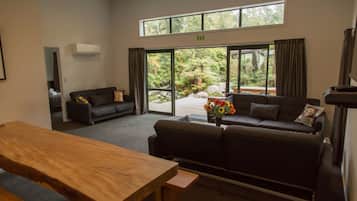 Uma sala de estar moderna com uma mesa de madeira, um sofá e um divã. Há uma janela grande com cortinas, um aparelho de ar condicionado e uma placa indicando uma saída.