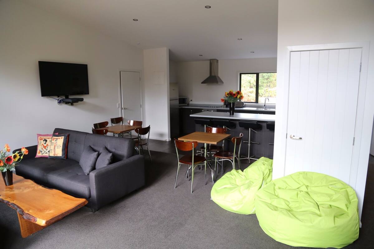 Una sala de estar moderna con un sofá, una mesa de centro y pufs de color verde. Se ve un comedor con una mesa y sillas. En el fondo, una cocina con estufa y fregadero.