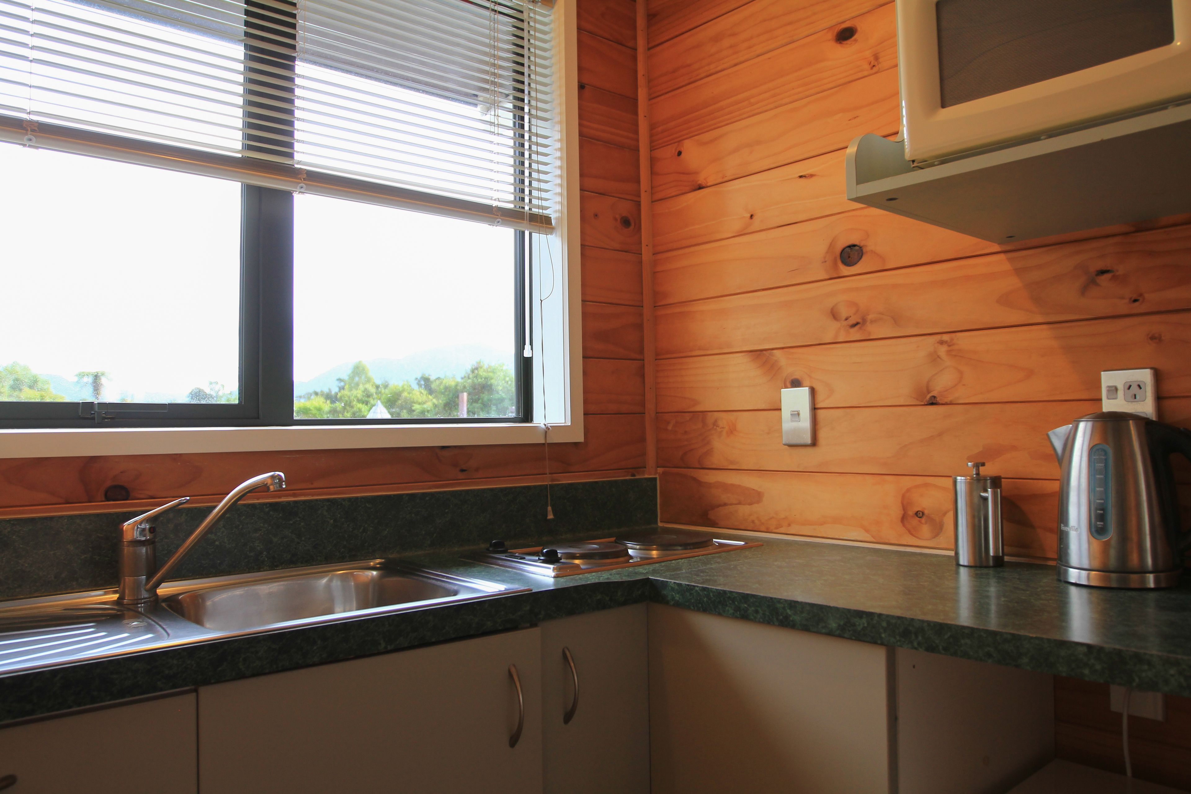 A kitchen with wooden walls, a window with blinds, a stainless steel sink, and a kettle on the counter.