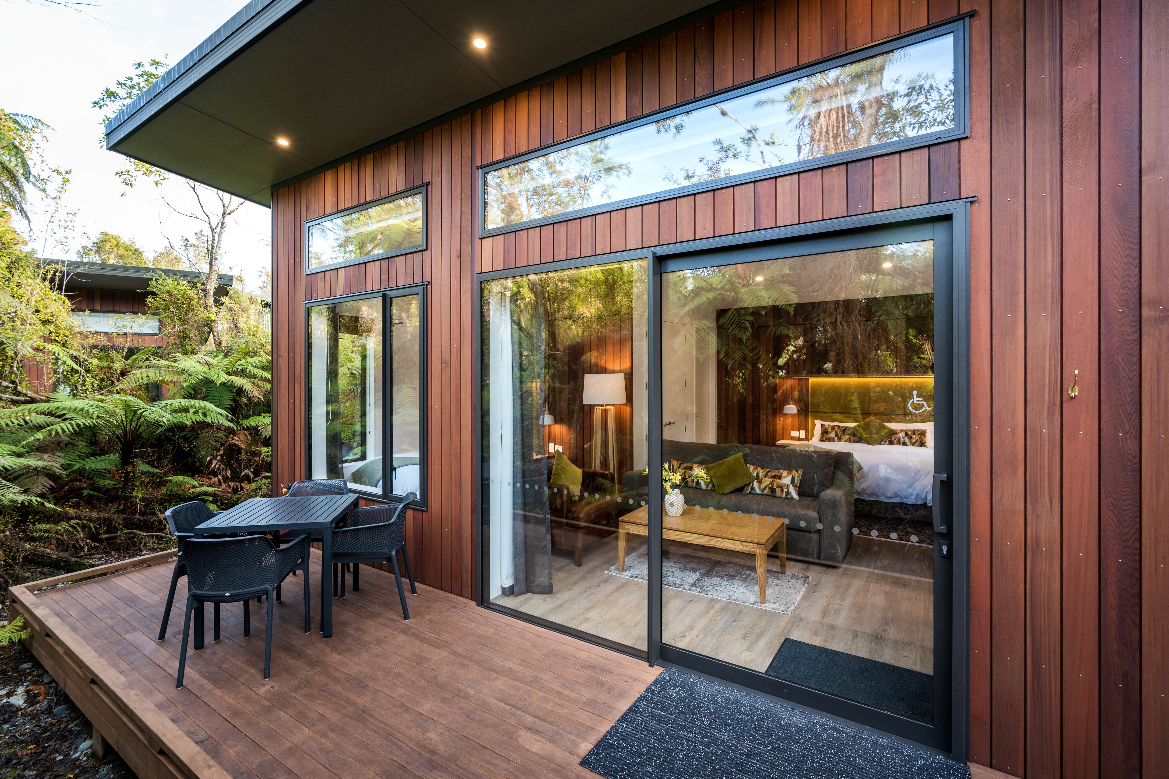 A modern house with a deck, large sliding glass doors, and a visible interior with a bed, sofa, and table.