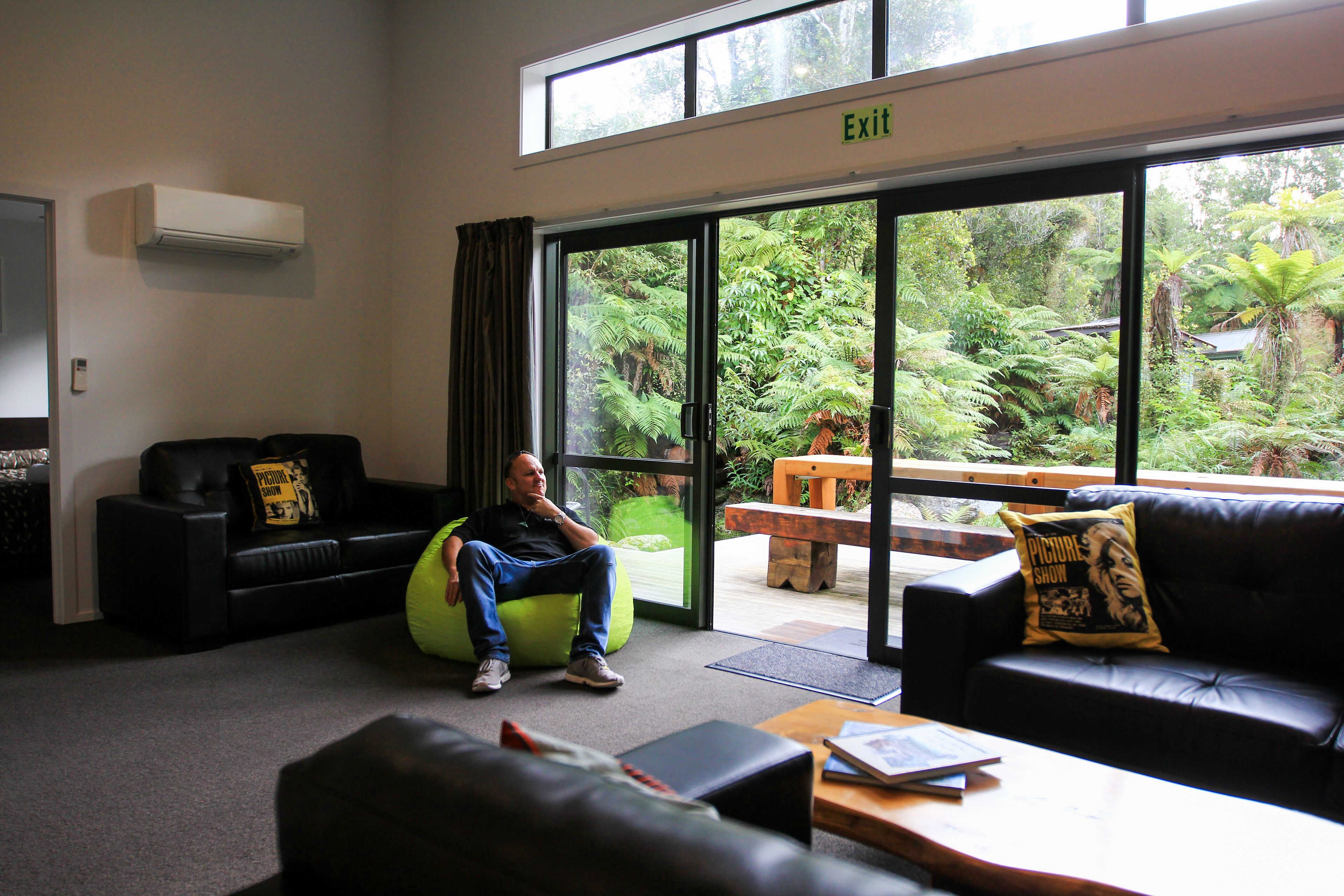 A modern interior with a black leather sofa, a green bean bag chair, and a wooden coffee table. A large window offers a view of lush greenery outside.