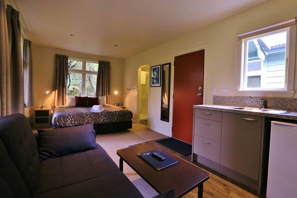 Una habitación de hotel compacta con cama, sofá, una mesita, kitchenette y ventana.