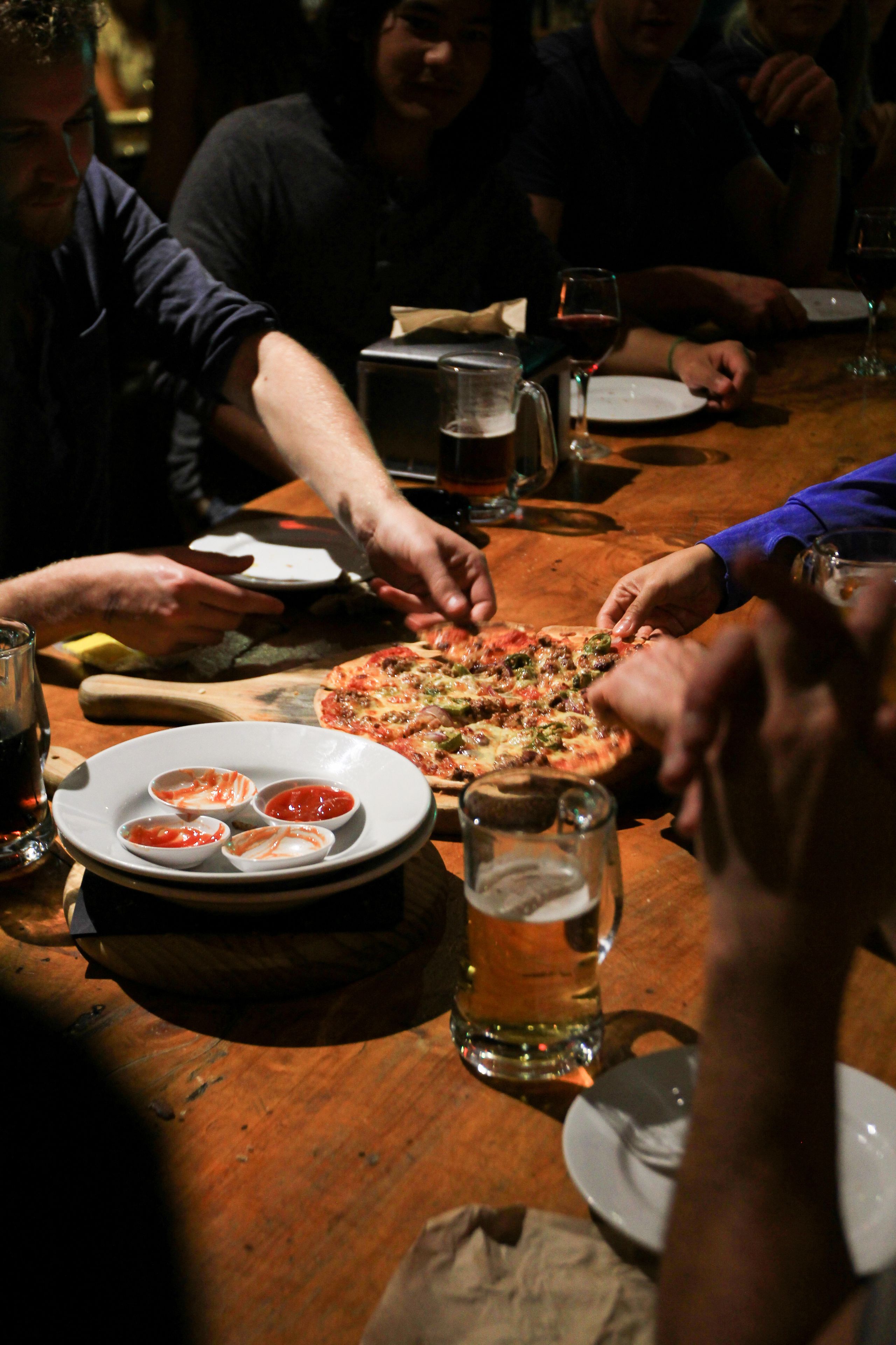 Un grupo de personas alrededor de una mesa de madera con pizza, vasos de cerveza y pequeños platos de acompañamientos.