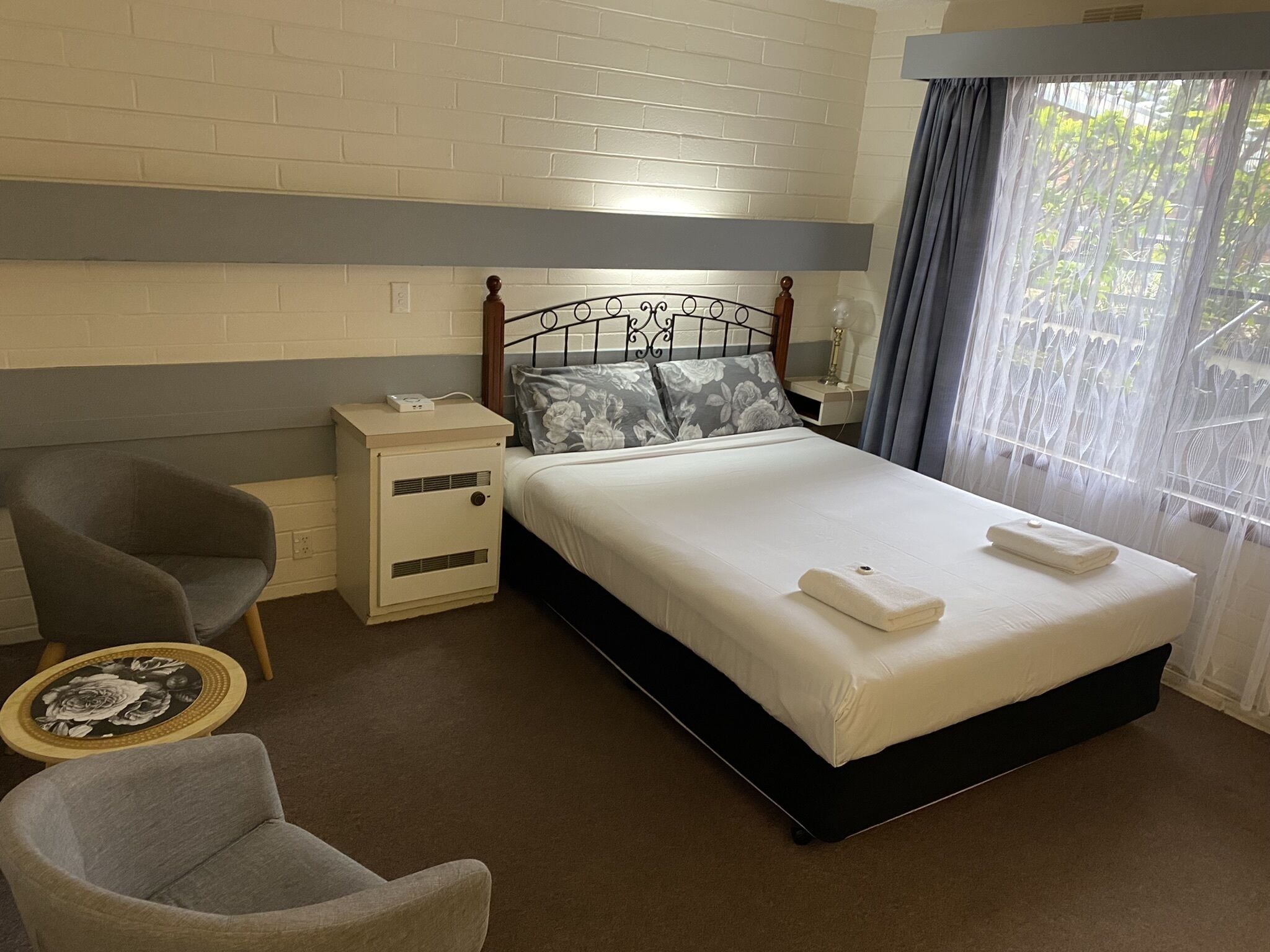 A hotel room with a bed, two chairs, a bedside table, and a window with curtains.