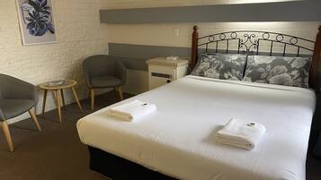 A neatly made bed with two towels, a grey armchair, a small round table, and a nightstand with a lamp.