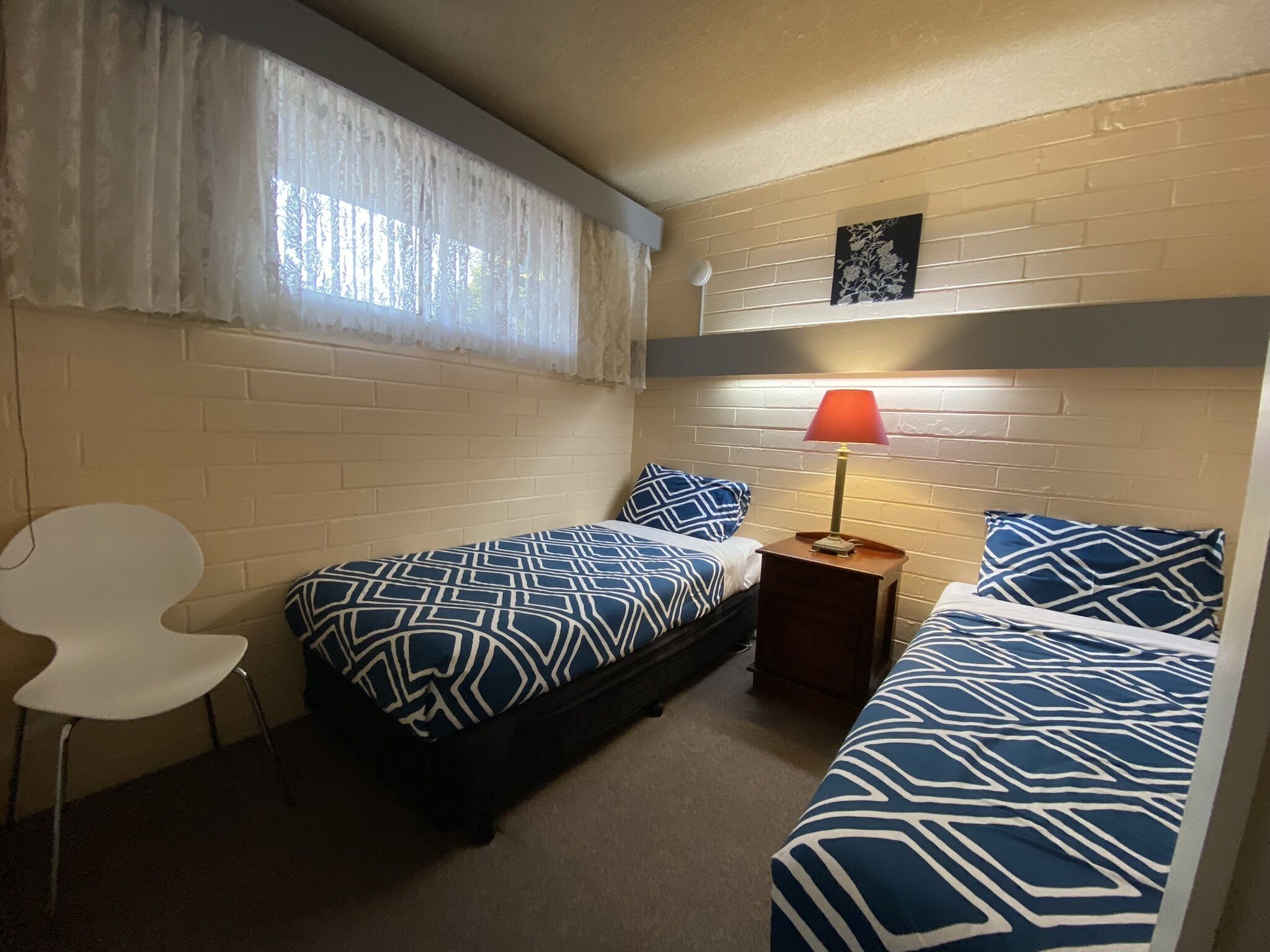 Una habitación pequeña y acogedora con dos camas, una silla, una mesita de noche con lámpara y una ventana con cortinas traslúcidas.