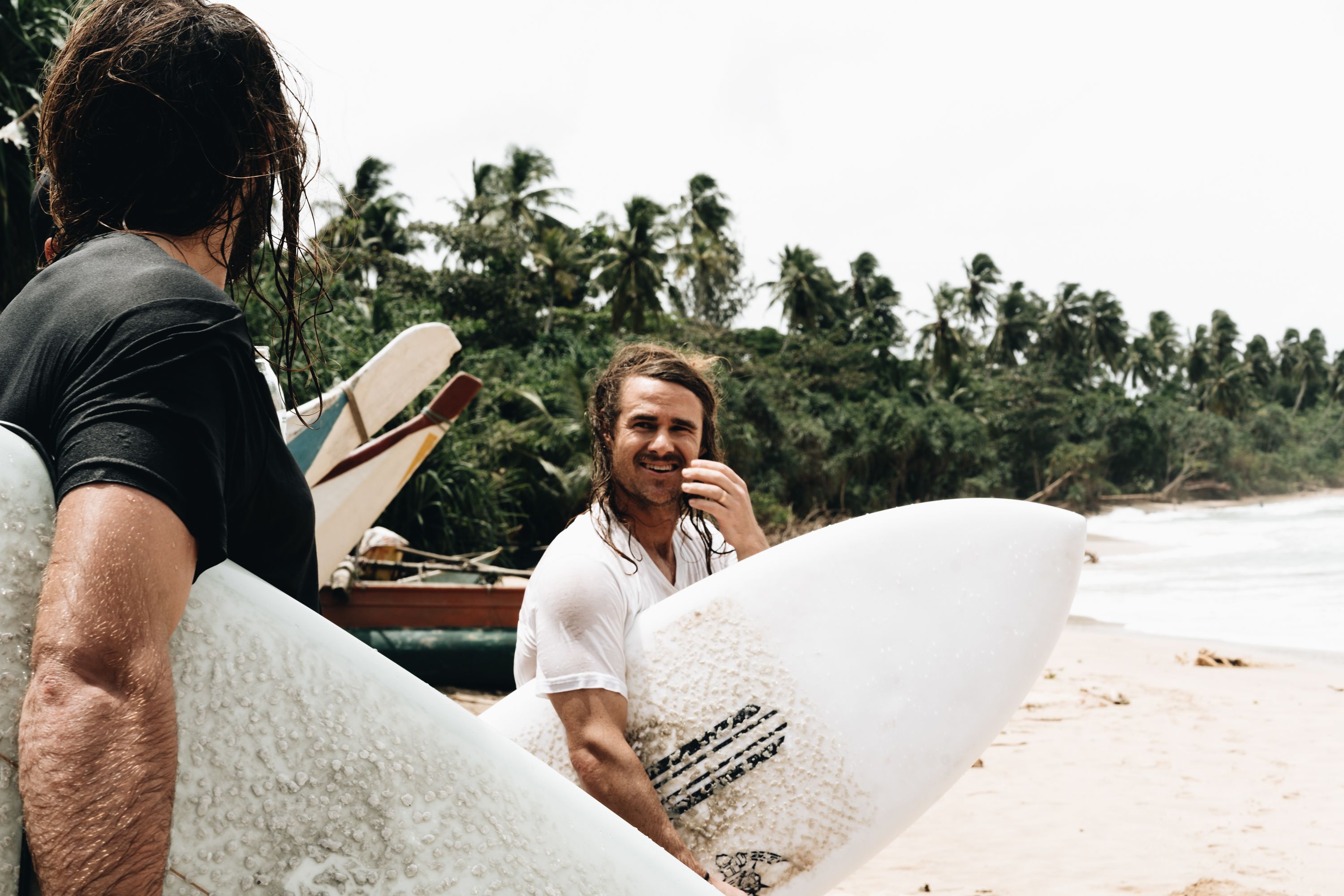 Two people holding surfboards on a beach with palm trees in the background.