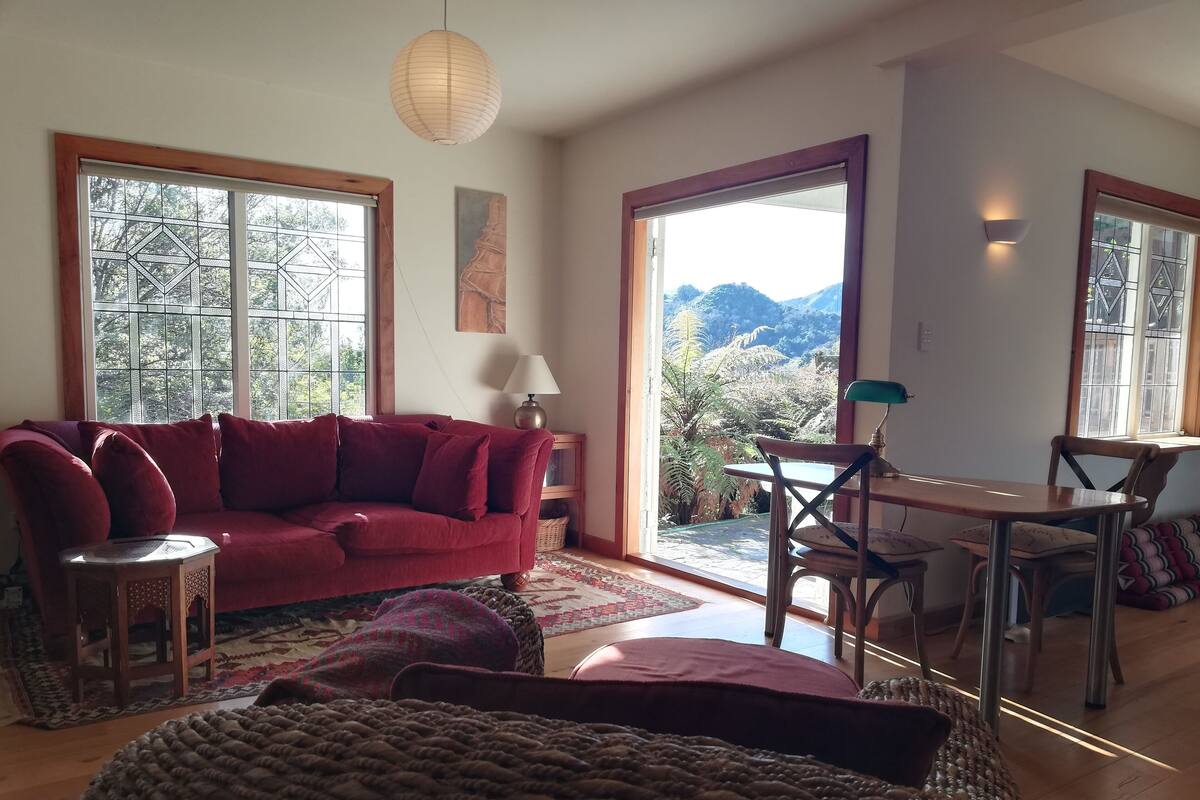 Ein gemütliches Wohnzimmer mit einem roten Sofa, einem Holzespeisetisch mit Stühlen und großen Fenstern mit Blick ins Freie.