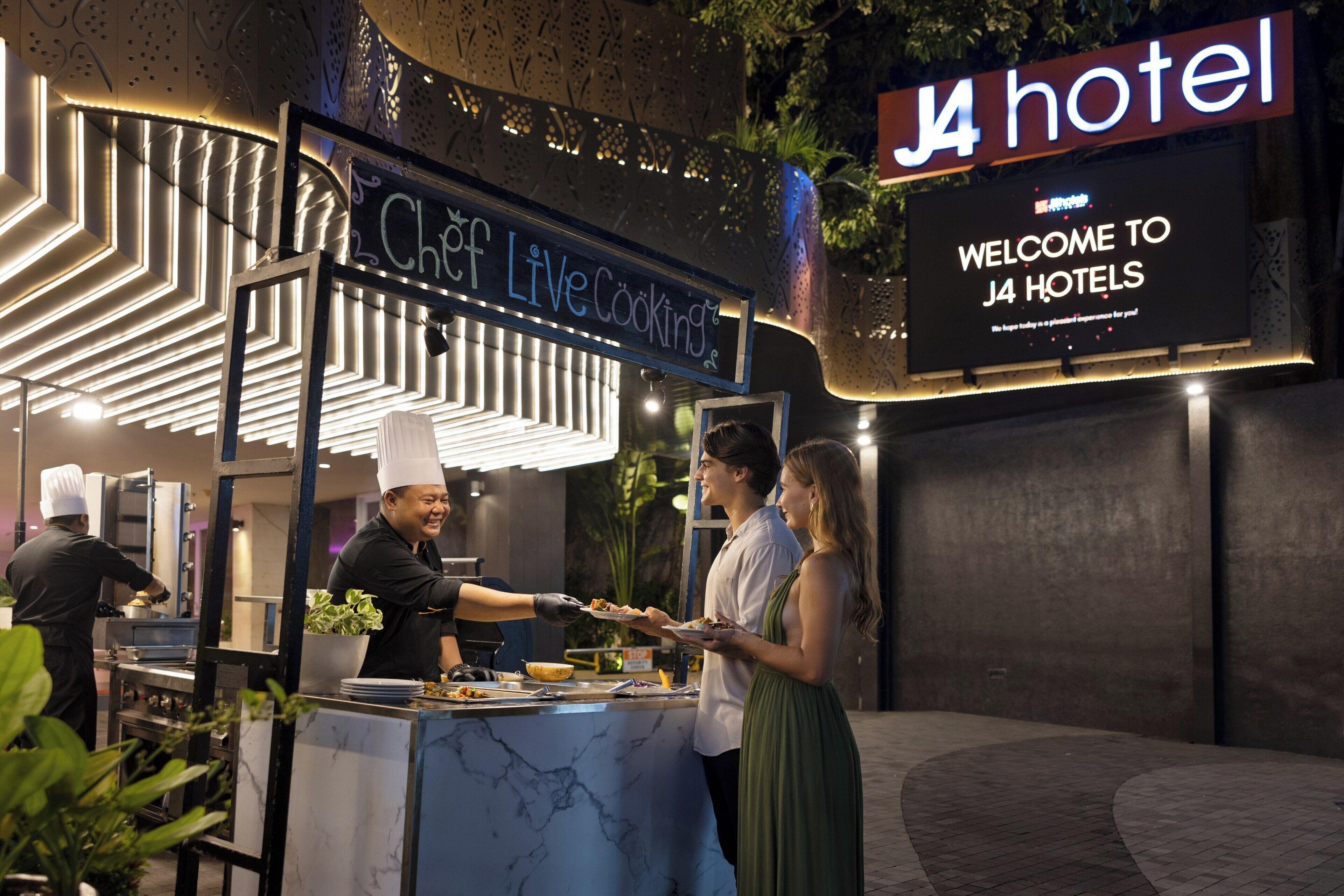 A chef serving food at a live cooking station with a sign for J4 hotel and a welcome sign.