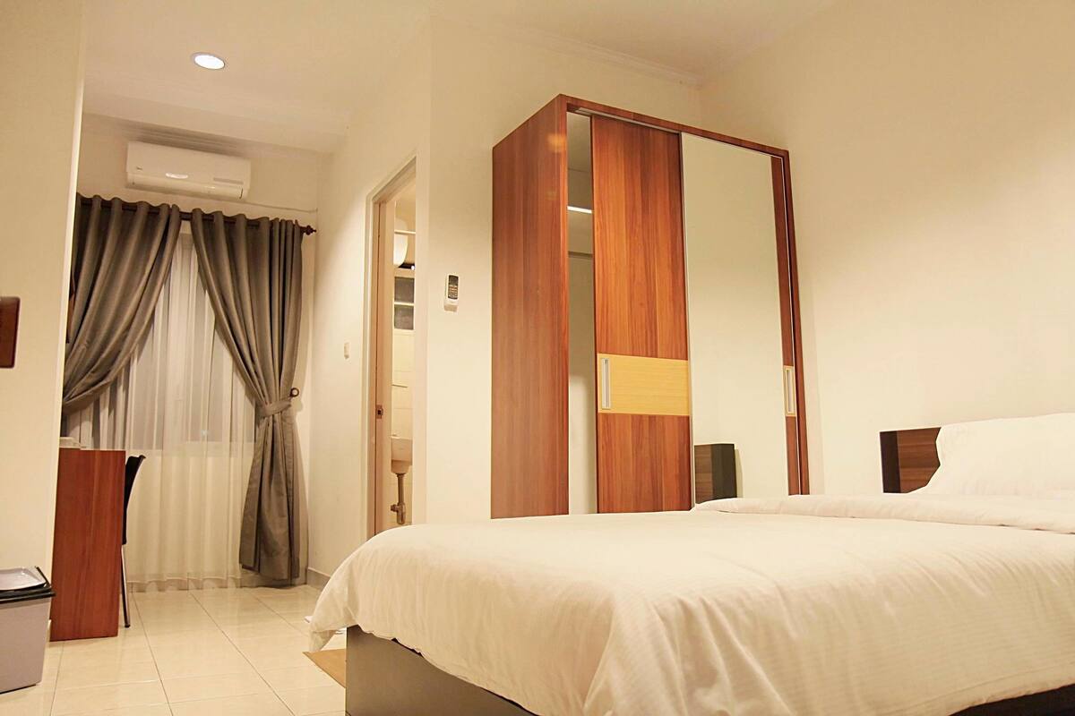A hotel room with a bed, a wardrobe, a mirror, a window with curtains, and an air conditioning unit.