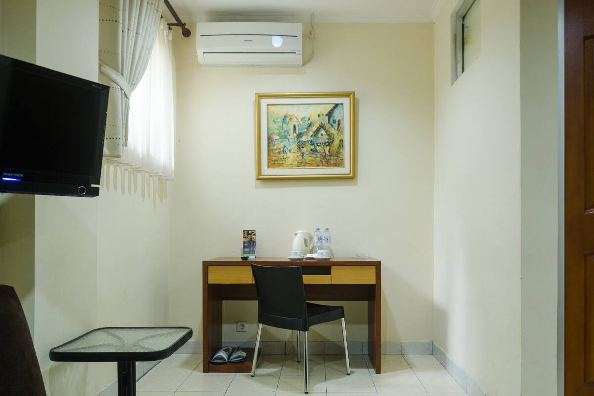 A small room with a wooden desk, a black chair, a television, a painting on the wall, and a small table with a chair.