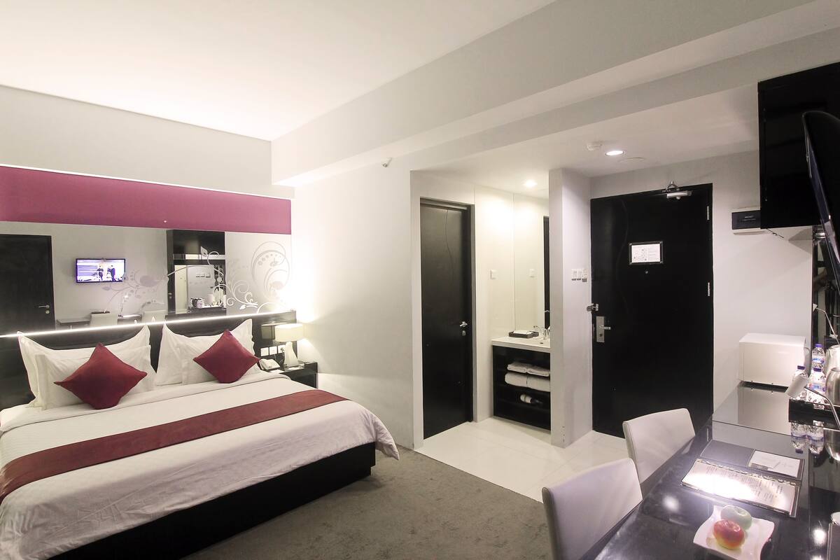 Una habitación de hotel moderna con una cama grande, un escritorio con silla, un televisor de pantalla plana y un baño con espejo y lavamanos.