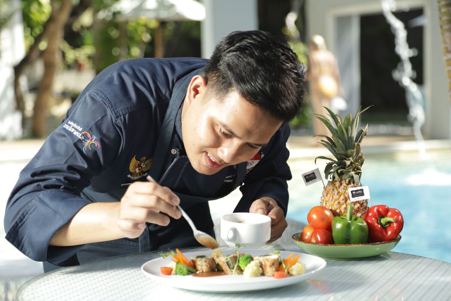 A person dining at a table by a poolside with a plate of food, a pineapple, and a bowl of vegetables.