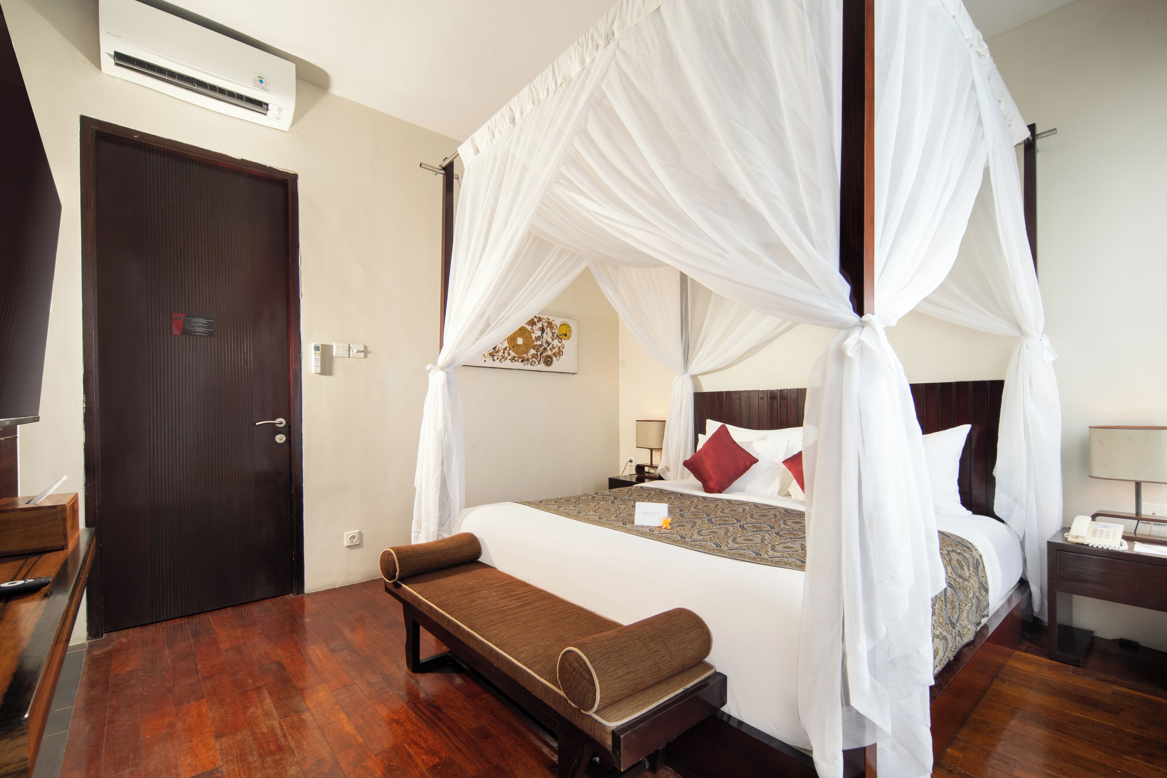 A hotel room with a four-poster bed, a bench, a desk with a telephone, and a wall-mounted air conditioner.