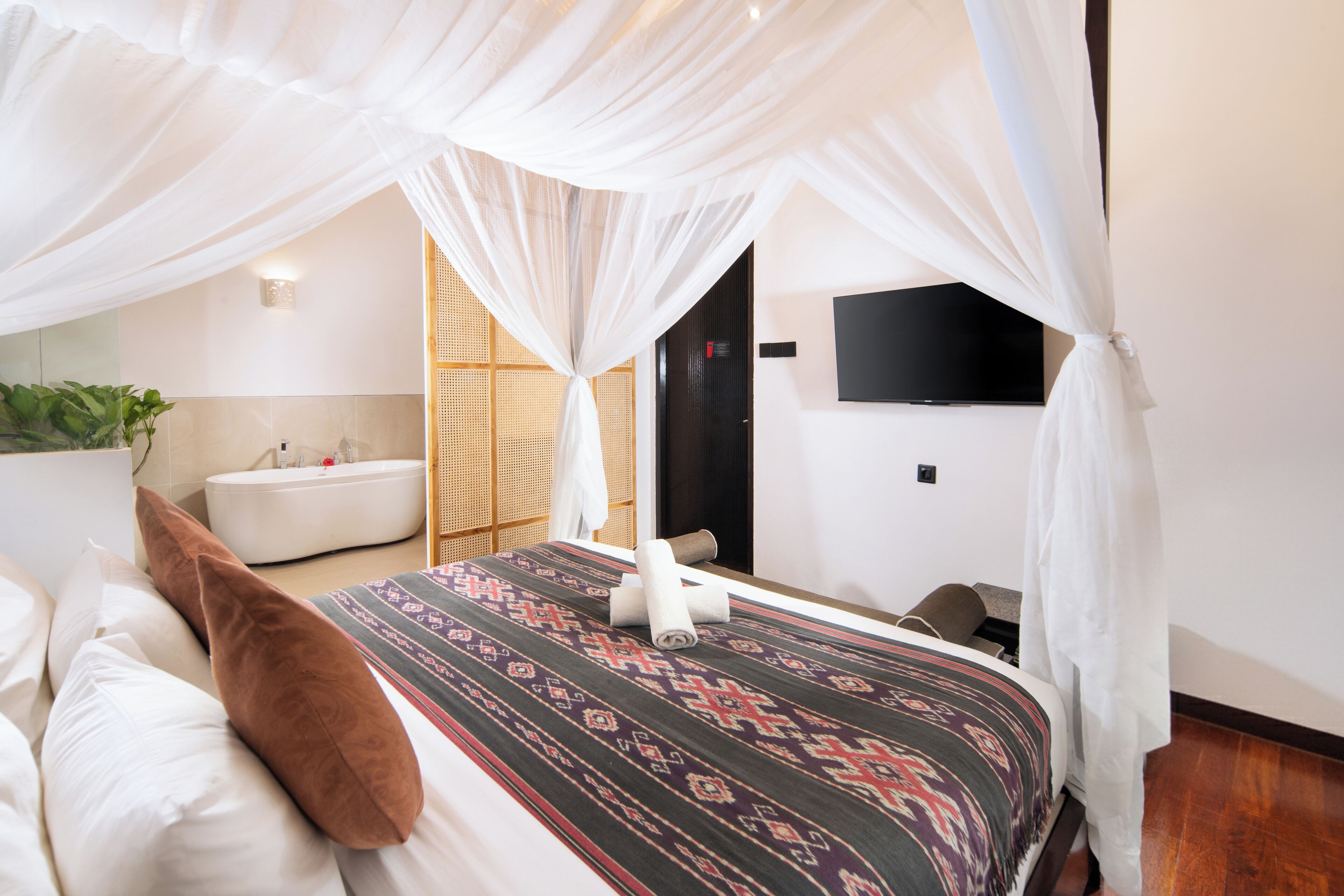 A bedroom with a bed, a white canopy, a TV on the wall, and a bathroom with a bathtub.
