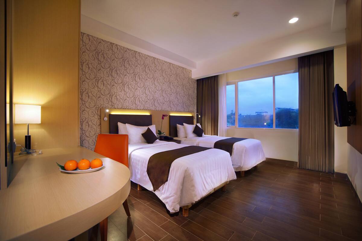Une chambre d’hôtel avec deux lits, une table à manger avec des oranges, une télévision et une grande fenêtre donnant sur un beau paysage.