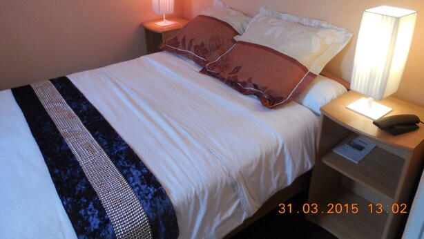A neatly made bed with white and brown bedding, two bedside lamps, a nightstand with a phone, and a visible date stamp. - Hôtel Le Petit Château (Malakoff)