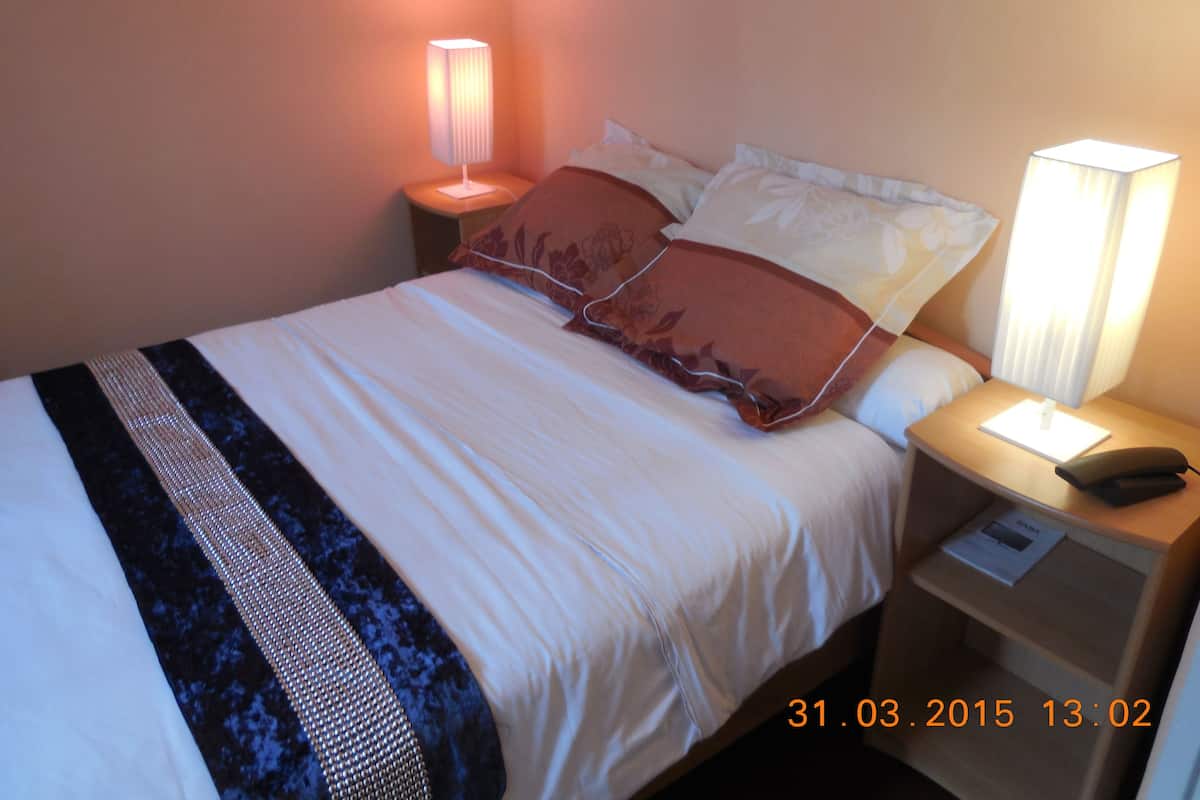 A neatly made bed with white and brown bedding, two bedside lamps, a nightstand with a phone, and a visible date stamp.