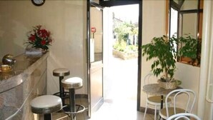 A hotel lobby with a reception desk, two bar stools, a potted plant, and a view of the outside. - Hôtel Le Petit Château (Malakoff)
